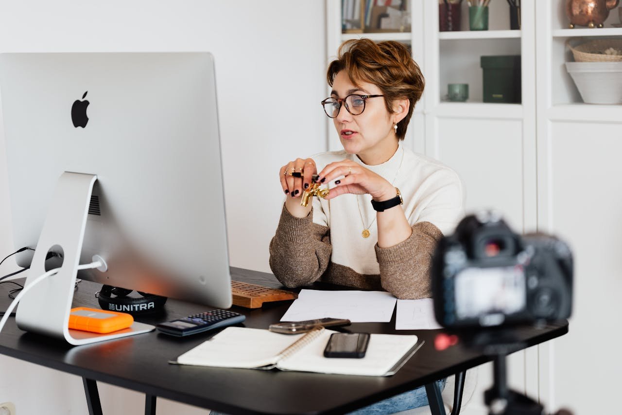 about-01 A professional woman engaged in a virtual meeting setup at her home desk, using a computer and camera.